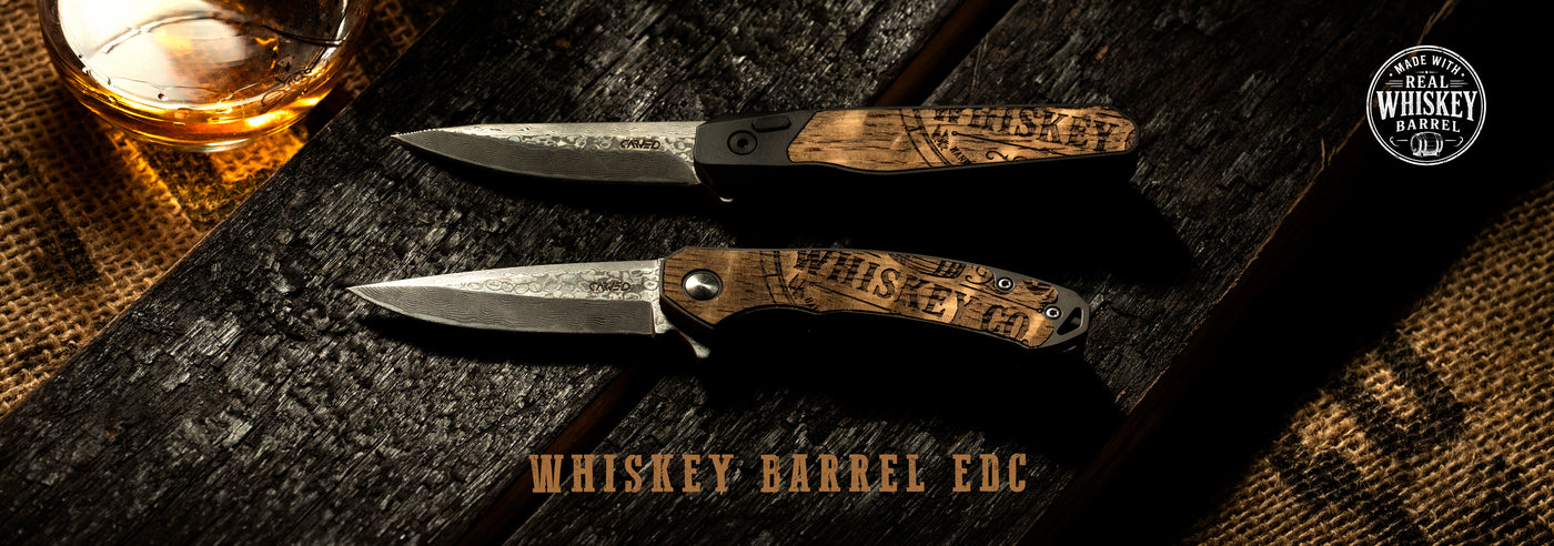Two Whiskey Barrel EDC knives on charred wood beside a glass of whiskey, with a made-with-real-whiskey-barrel badge.